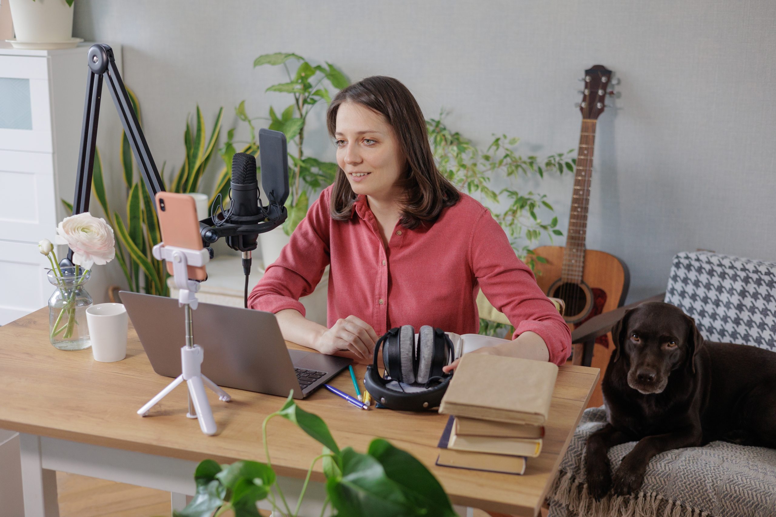 woman in a home office records podcast audio content with a microphone and headphones and a labrador dog